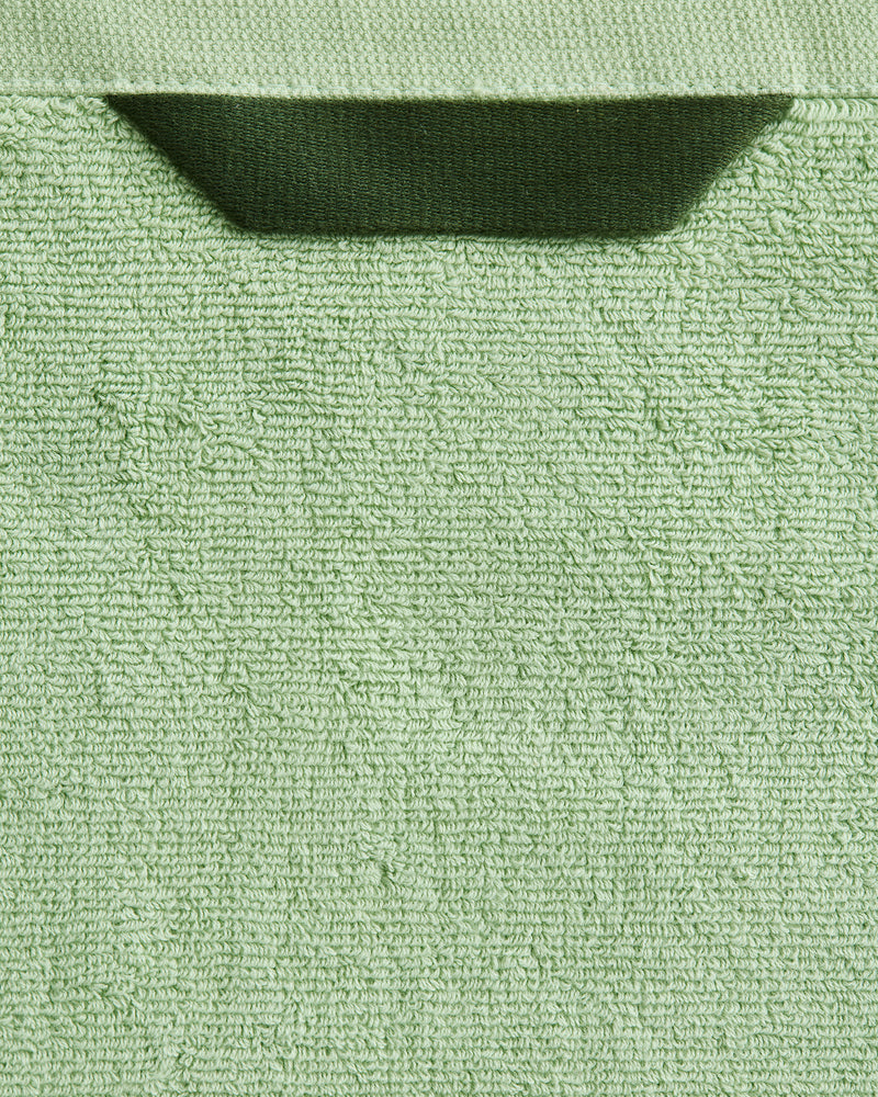 A close-up of the Bed Threads Apple 100% Cotton Terry Bath Sheet Bundle in light green, featuring a dark green fabric loop at the top edge, set against a matching green background.