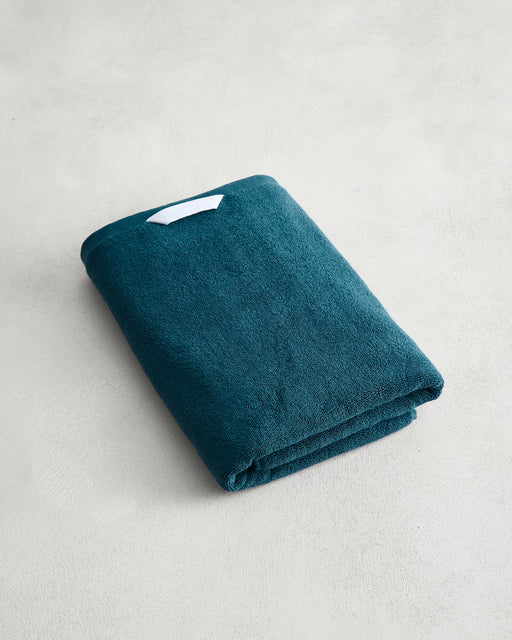 The Aegean 100% Cotton Terry Bath Towel by Bed Threads, a teal 650GSM towel with a small white tag, rests on a light gray textured surface and offers quick-drying convenience.