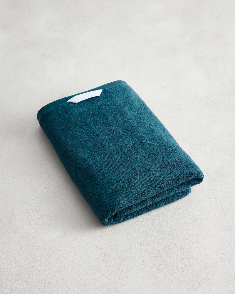 The Aegean 100% Cotton Terry Bath Towel by Bed Threads, a teal 650GSM towel with a small white tag, rests on a light gray textured surface and offers quick-drying convenience.