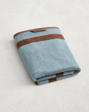 The Bed Threads Cacao & Lagoon Two-Toned 100% Cotton Terry Bath Towel, featuring blue and brown stripes plus a small brown tag, is folded on a light textured surface. - view