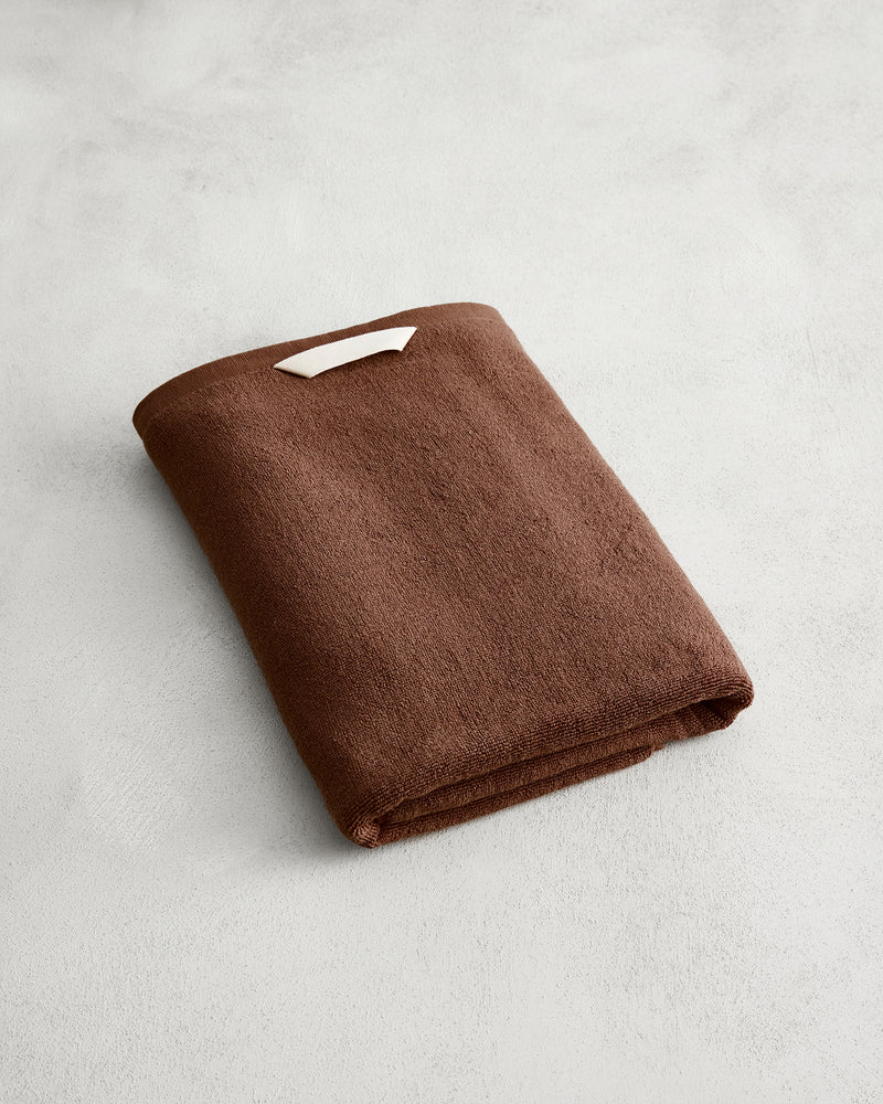 The Bed Threads Espresso 100% Cotton Terry Bath Towel, neatly folded with a small white label, rests on a light gray textured surface.