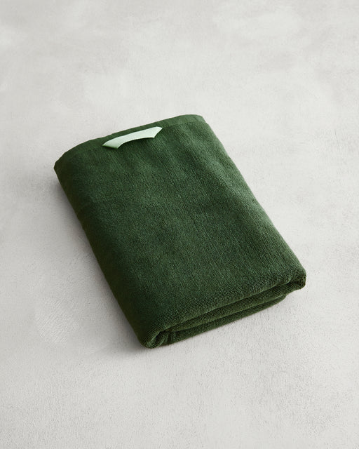 A folded Moss 100% Cotton Terry Bath Towel by Bed Threads in dark green rests on a light grey textured surface, with a small light green tag attached to its corner.