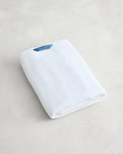 The Crisp White 100% Cotton Terry Bath Towel by Bed Threads, neatly folded with a small blue tag, rests on a light, textured surface. - view