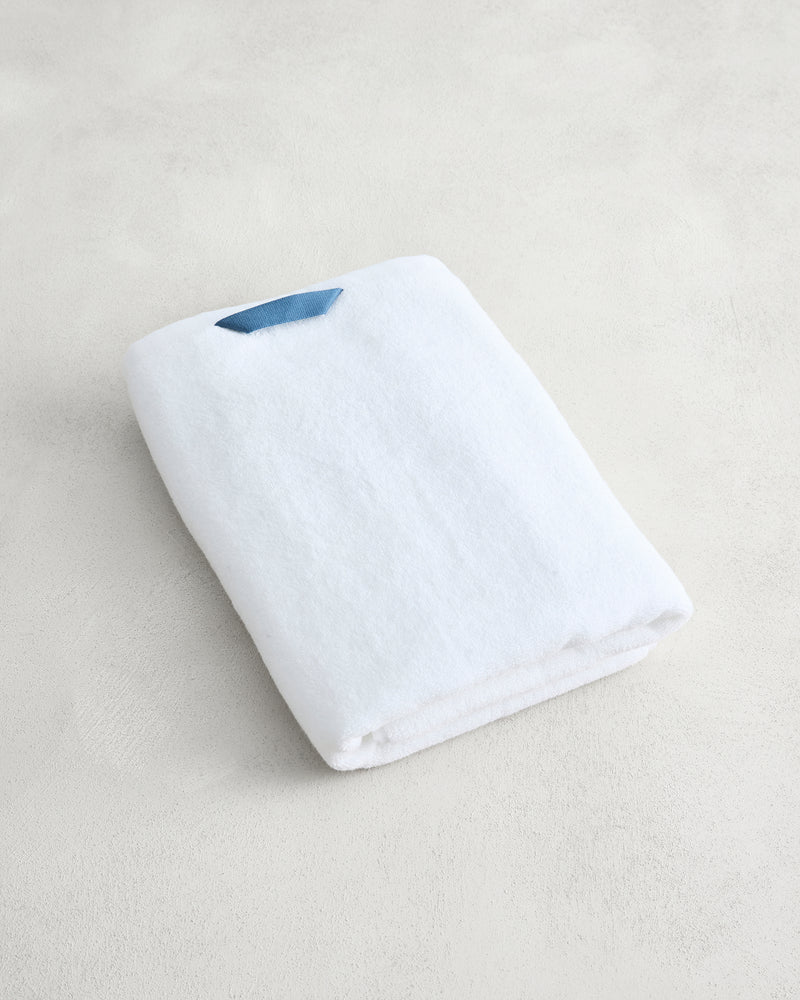 The Crisp White 100% Cotton Terry Bath Towel by Bed Threads, neatly folded with a small blue tag, rests on a light, textured surface.