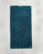 The Aegean 100% Cotton Terry Bath Towel by Bed Threads, a dark teal rectangular towel with a small white tag, crafted from 650GSM cotton, is displayed on a light gray background. - view