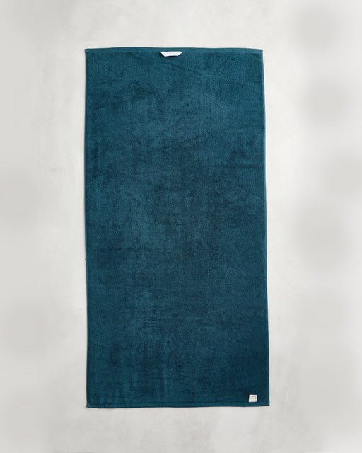 The Aegean 100% Cotton Terry Bath Towel by Bed Threads, a dark teal rectangular towel with a small white tag, crafted from 650GSM cotton, is displayed on a light gray background.