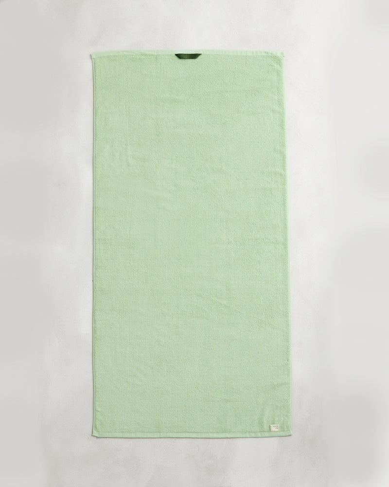 A set of two Apple 100% Cotton Terry Bath Towels by Bed Threads, in light green, lies flat on a white surface, each featuring a small fabric tag in the corner and a hanging loop at the top edge.