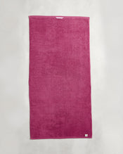 A rectangular Berry 100% Cotton Terry Bath Towel by Bed Threads hangs flat against a light gray wall. The plush magenta towel has a small white tag on the bottom right corner. - view