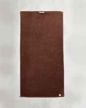 An Espresso 100% Cotton Terry Bath Towel by Bed Threads is laid flat on a light gray surface, featuring a small white label on the bottom corner and a beige hanging loop near the top edge. - view