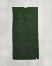 A Moss 100% Cotton Terry Bath Towel from Bed Threads hangs vertically against a white background, displaying a light blue tag at the top and a small label in the bottom right corner. - view