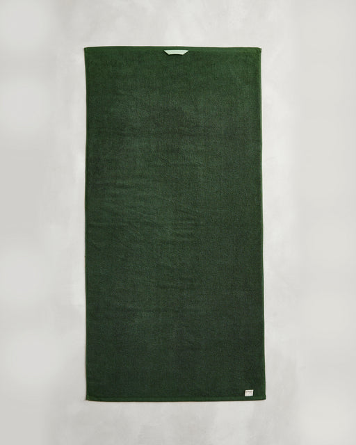 A Moss 100% Cotton Terry Bath Towel from Bed Threads hangs vertically against a white background, displaying a light blue tag at the top and a small label in the bottom right corner.