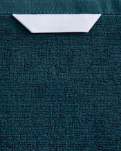 A white paper boat rests near the top center on a textured dark teal surface, evoking the plush feel of Bed Threads' Aegean 100% Cotton Terry Bath Towel. - view