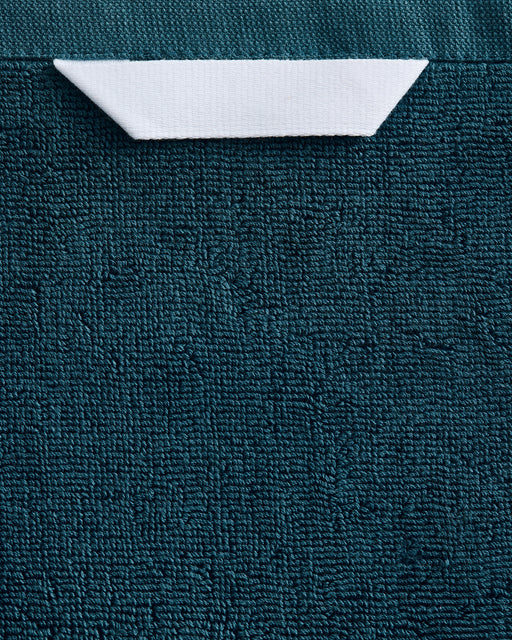 A white paper boat rests near the top center on a textured dark teal surface, evoking the plush feel of Bed Threads' Aegean 100% Cotton Terry Bath Towel.