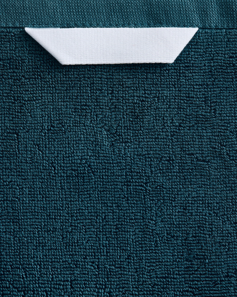 A white paper boat rests near the top center on a textured dark teal surface, evoking the plush feel of Bed Threads' Aegean 100% Cotton Terry Bath Towel.