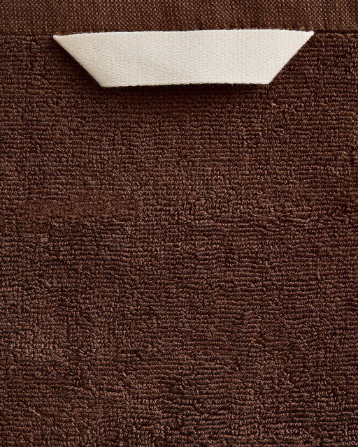 A close-up of the Bed Threads Espresso 100% Cotton Terry Bath Towel, showing its brown textured surface with a white triangular corner folded down to reveal the reverse side.