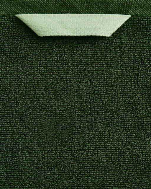 A close-up of the Bed Threads Moss 100% Cotton Terry Bath Towel Bundle shows its dark green textured surface, with a light green corner folded over to reveal the smooth backside and contrast with the highly absorbent terry fabric.