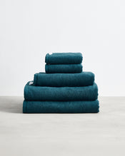 A stack of four Aegean 100% Cotton Terry Bath Towels by Bed Threads in dark teal rests neatly folded on a light surface against a plain white background. - view