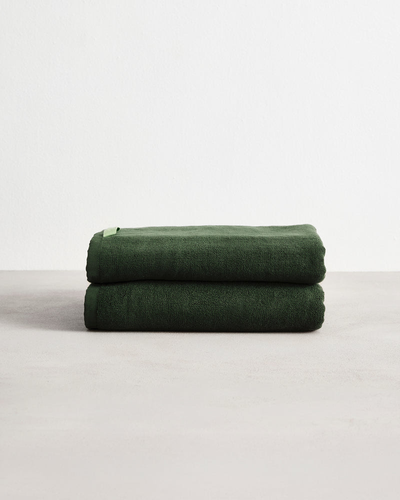 Two Moss 100% Cotton Terry Bath Towels (Set of Two) by Bed Threads, neatly folded in dark green, are stacked on a light surface with a plain white background.