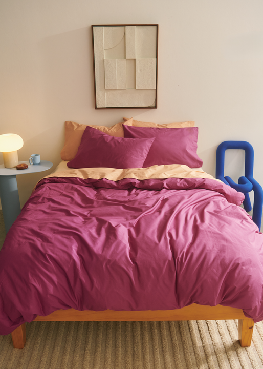 A bed with a magenta and peach reversible duvet and matching pillowcases on a wooden frame. Beside it sits a blue sculptural chair and a round side table with a lamp. Abstract wall art hangs above.