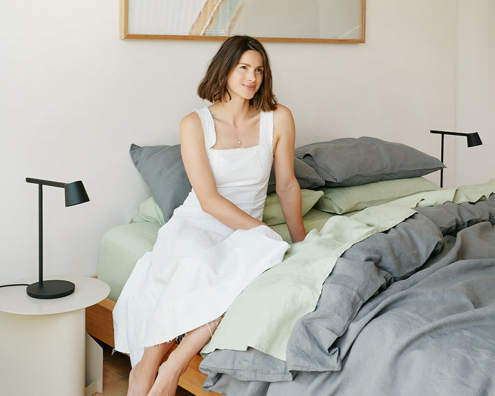A woman in a white dress sits on the edge of a bed with gray and light green bedding, in a bright, modern bedroom with minimalist decor and black bedside lamps.