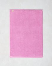 The Bubblegum 100% Cotton Terry Bath Mat by Bed Threads, in a rectangular pink design, lies flat on a light textured surface with a small fabric tag visible on one corner. - view