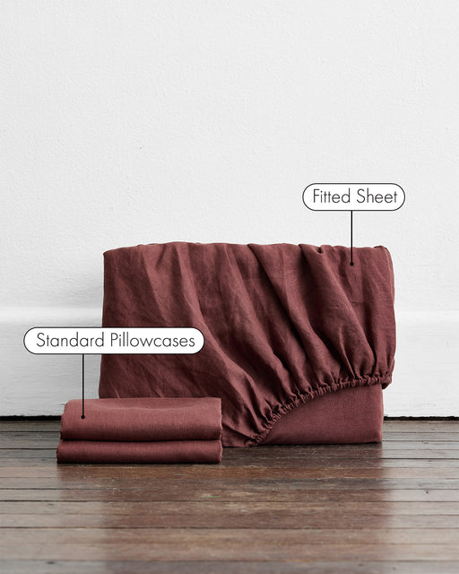 A folded stack of Bed Threads' Cacao 100% French Flax Linen Fitted Sheet Set lies on a wooden floor, with two “Standard Pillowcases” and a neatly folded fitted sheet displaying its elastic edge.