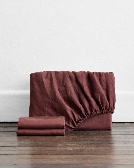 The Bed Threads Cacao 100% French Flax Linen Fitted Sheet Set, a warm reddish-brown, features both flat and fitted sheets—perfect for enhancing your linen bedding collection. Displayed neatly on a wooden floor against a white wall.