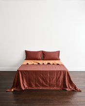 A bed dressed in a rust brown quilt cover in Cacao & Hazelnut with two matching pillowcases in Cacao, set against a plain white wall and dark wood floor. - view