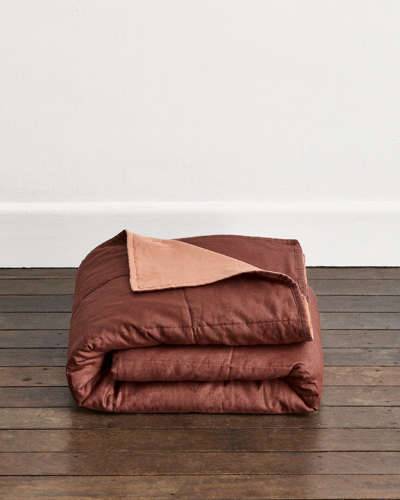 A folded brown quilt cover in Cacao & Hazelnut with or a Hazelnut underside, displayed on a dark wooden floor against a plain white wall.