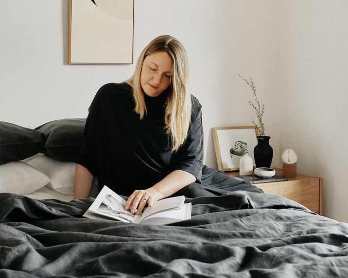 A woman with blonde hair sits on a bed with dark bedding, reading a book. Behind her, a nightstand holds framed photos, a vase with dried branches, and other decorative objects in a minimalist bedroom.