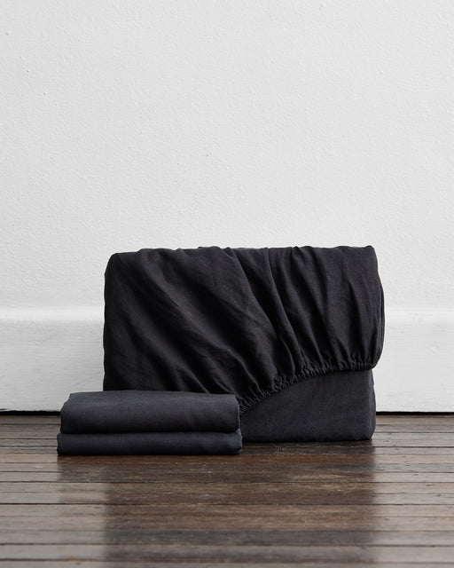 Two folded black linen sheets sit in front of a larger, unfolded Bed Threads Charcoal 100% French Flax Linen Fitted Sheet Set with elasticized edges on a dark wooden floor.
