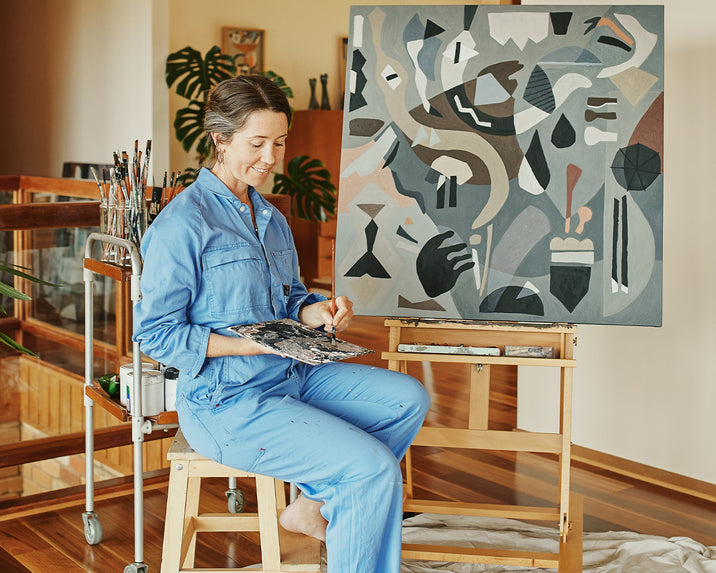 A woman in a blue jumpsuit sits on a stool, smiling as she paints an abstract artwork on a canvas. She holds a palette and brushes, with art supplies nearby in a bright, modern room.
