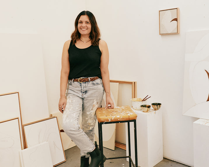 A woman wearing a black tank top and paint-splattered jeans stands smiling in an art studio, surrounded by framed minimalist artwork and painting supplies on a stool.
