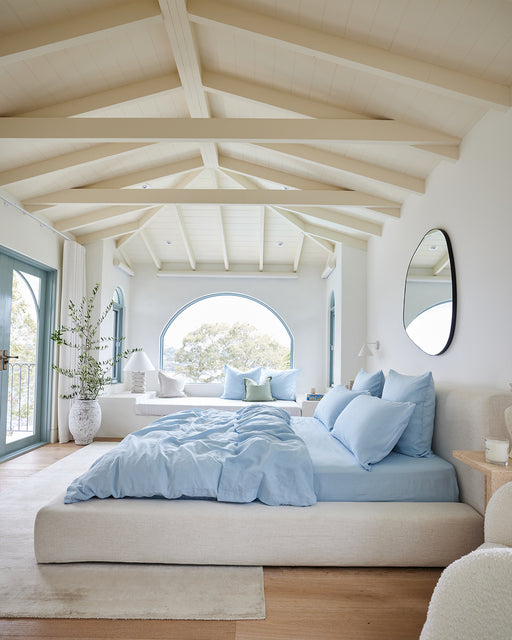 A bright, airy bedroom with high beamed ceilings, large arched windows, and light wood floors features a Bed Threads Coast 100% French Flax Linen Duvet Cover Set. Potted plants, a round mirror, and cozy seating complete the inviting space.