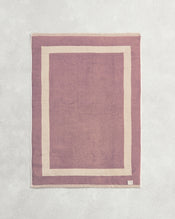 Flat cotton terry bath mat on a grey background in a lavender colour with a creme border. - view