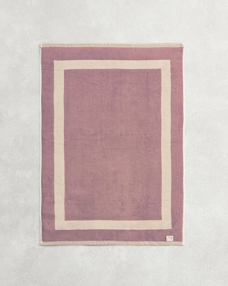 Flat cotton terry bath mat on a grey background in a lavender colour with a creme border.