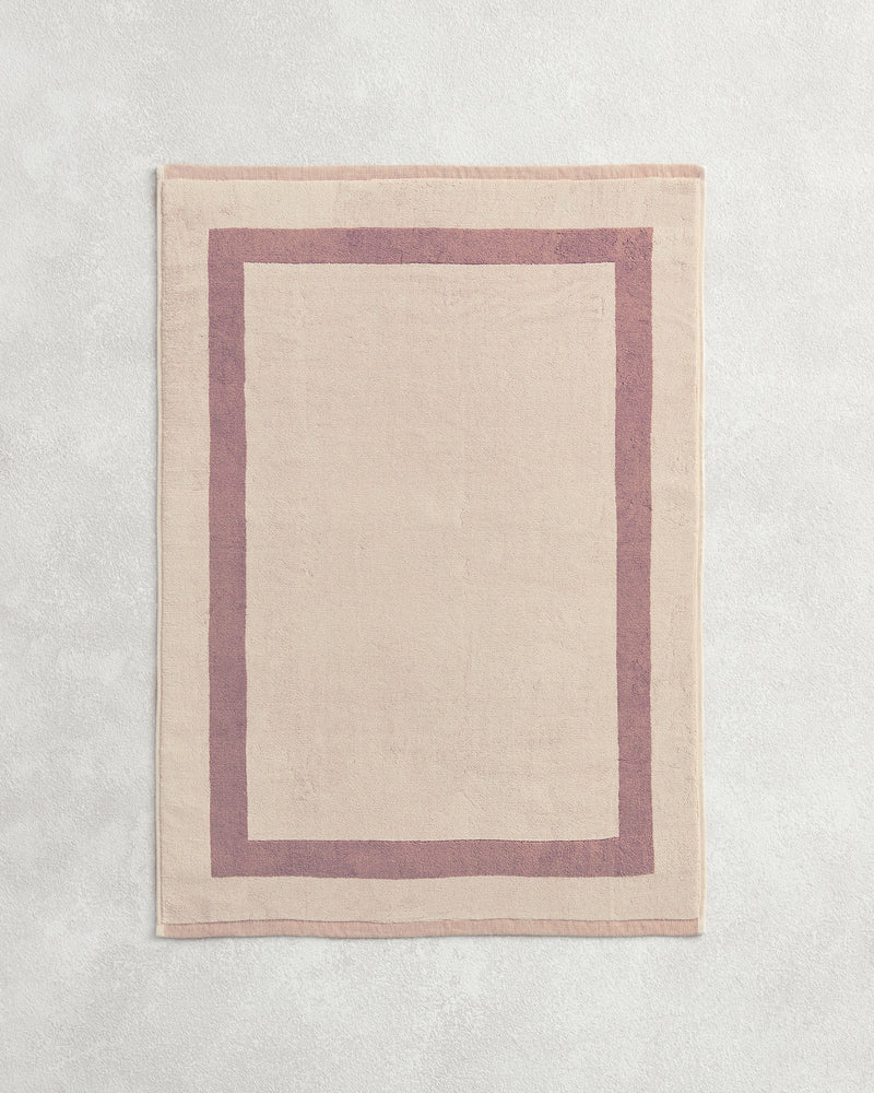 Flat cotton terry bath mat on a grey background in a creme colour with a lavender border.