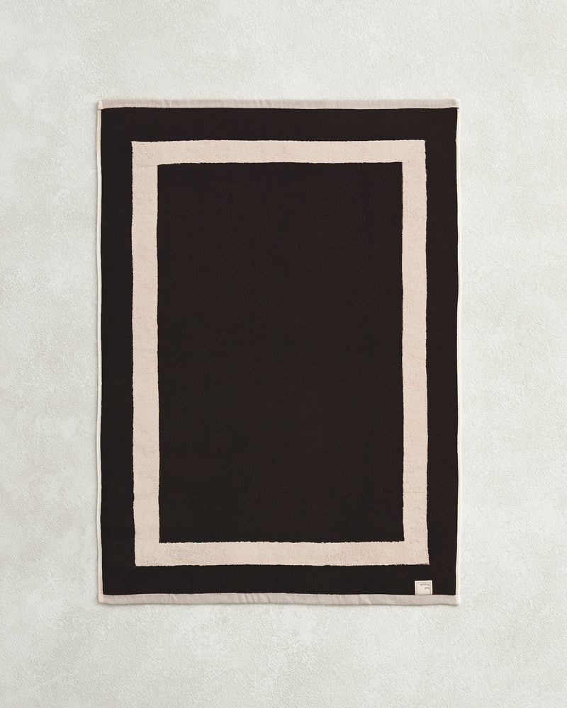 Flat cotton terry bath mat on a grey background in a charcoal colour with an oatmeal border.