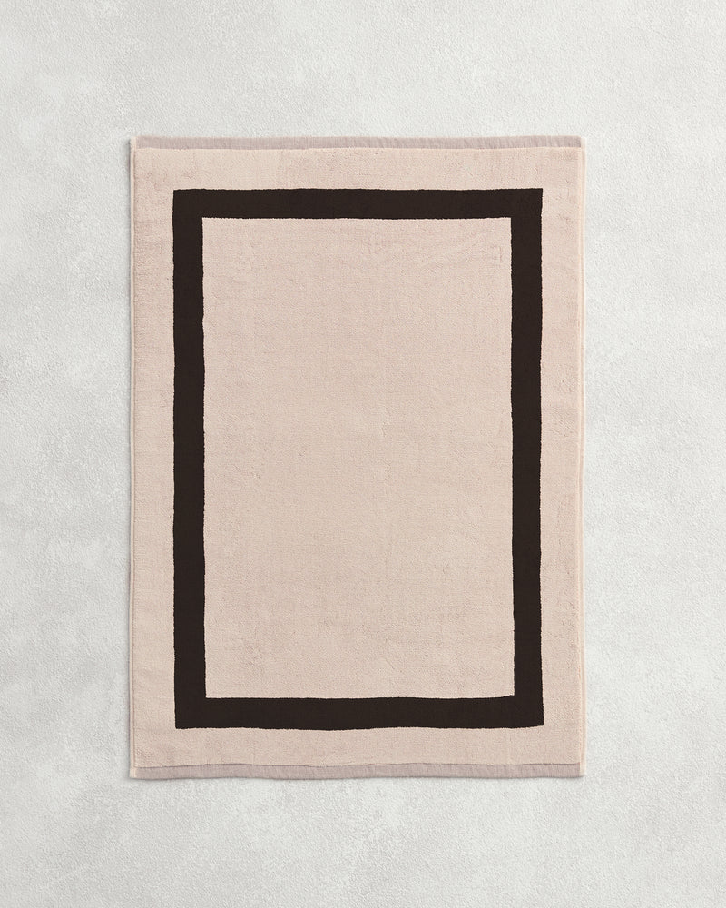 Flat cotton terry bath mat on a grey background in an oatmeal colour with a charcoal border.