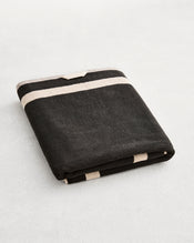 A folded cotton terry bath sheet on a grey background, in a charcoal colourway with an oatmeal border.  - view