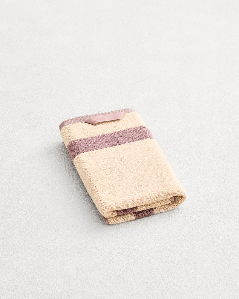 A folded cotton terry hand towel on a grey background, in a creme colourway with a lavender border. 