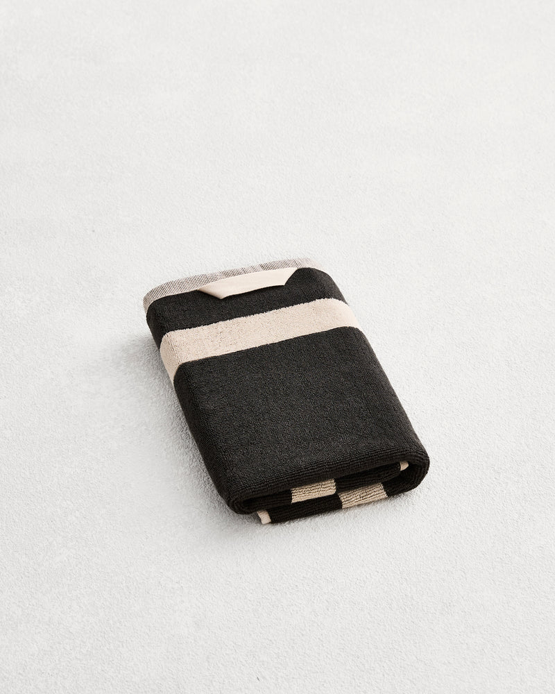 A folded cotton terry hand towel on a grey background, in a charcoal colourway with an oatmeal border. 
