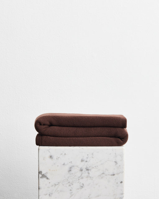 A folded dark brown wool cotton blanket in the shade mocha, stacked on top of a white marble pedestal against a plain white wall.