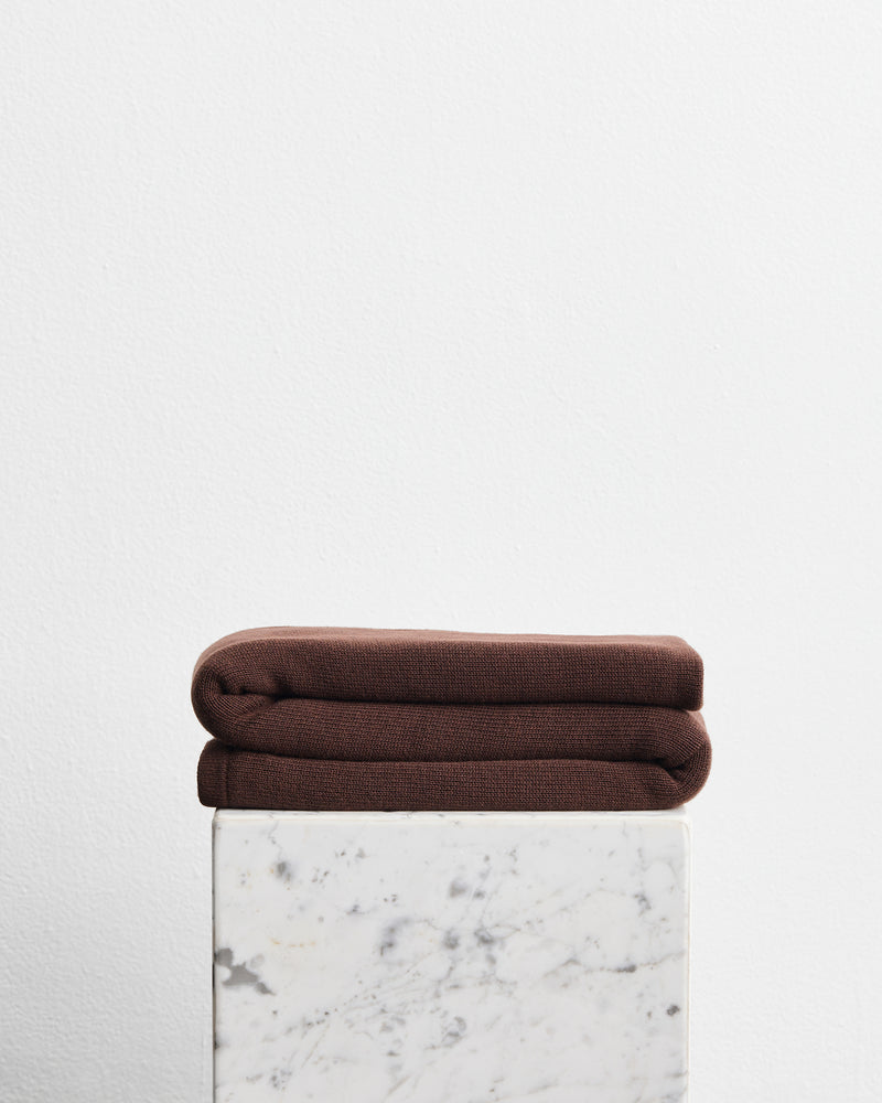 A folded dark brown wool cotton blanket in the shade mocha, stacked on top of a white marble pedestal against a plain white wall.