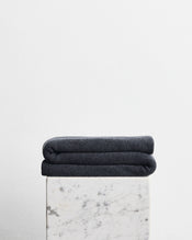 A folded dark charcoal gray wool cotton blanket in the shade shadow, stacked on top of a white marble pedestal against a plain white wall. - view
