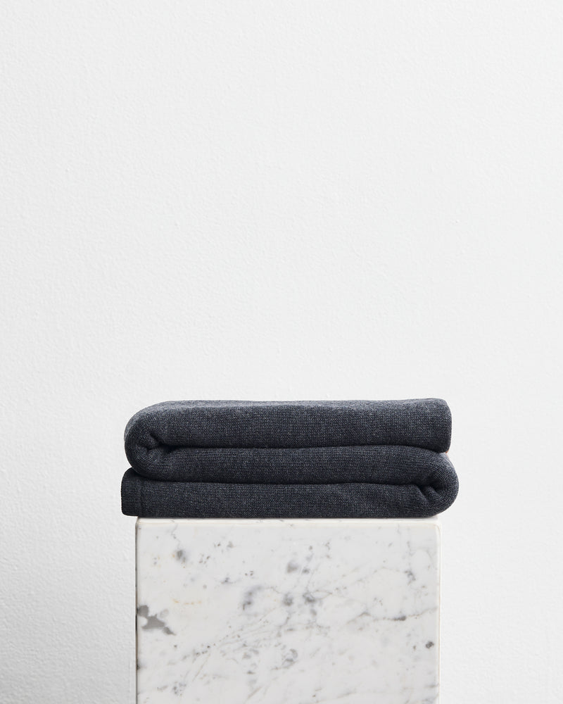 A folded dark charcoal gray wool cotton blanket in the shade shadow, stacked on top of a white marble pedestal against a plain white wall.