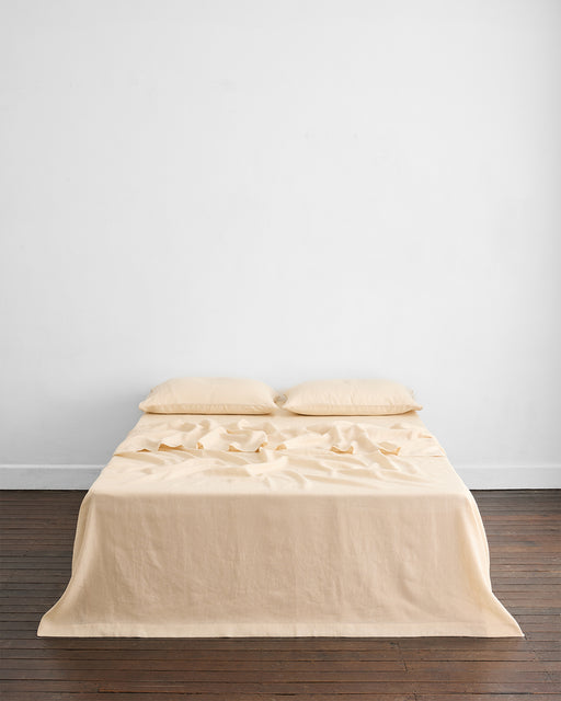 A neatly made bed with two Bed Threads Crème 100% French Flax Linen King Pillowcases rests against a plain white wall on a wooden floor. The slightly wrinkled linen bedding adds to the minimal, uncluttered look of the room.