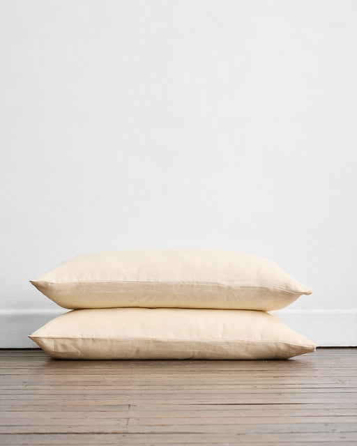 Two Crème 100% French Flax Linen King Pillowcases by Bed Threads are stacked on a wooden floor against a plain white wall, highlighting their simple and minimalistic design.