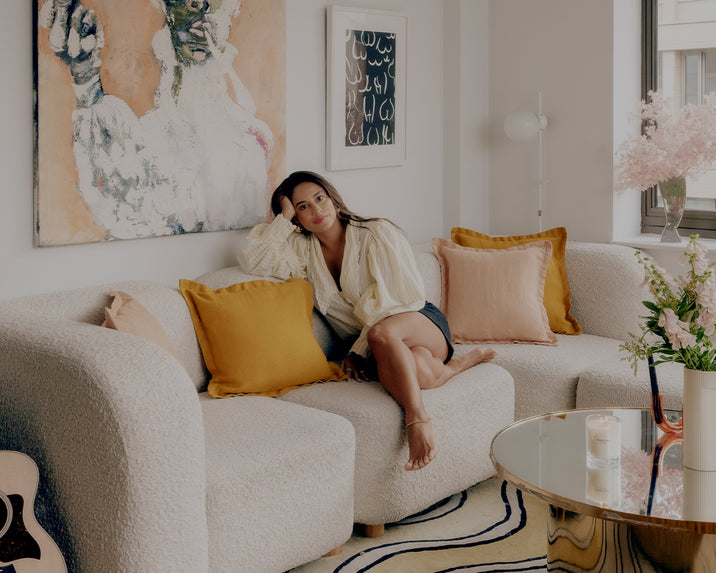 A woman sits on a textured white sofa with colorful cushions in a stylish living room, featuring wall art, a round glass coffee table, flowers, a candle, and natural light from a window.