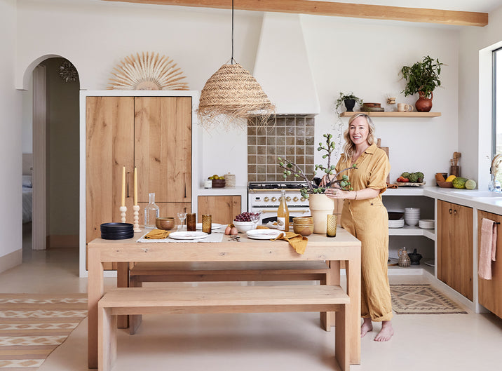 A smiling woman in a tan jumpsuit stands in a bright, modern kitchen with wood accents, setting a rustic dining table adorned with plates, glasses, and a branch in a vase. Green plants and natural light fill the space.
